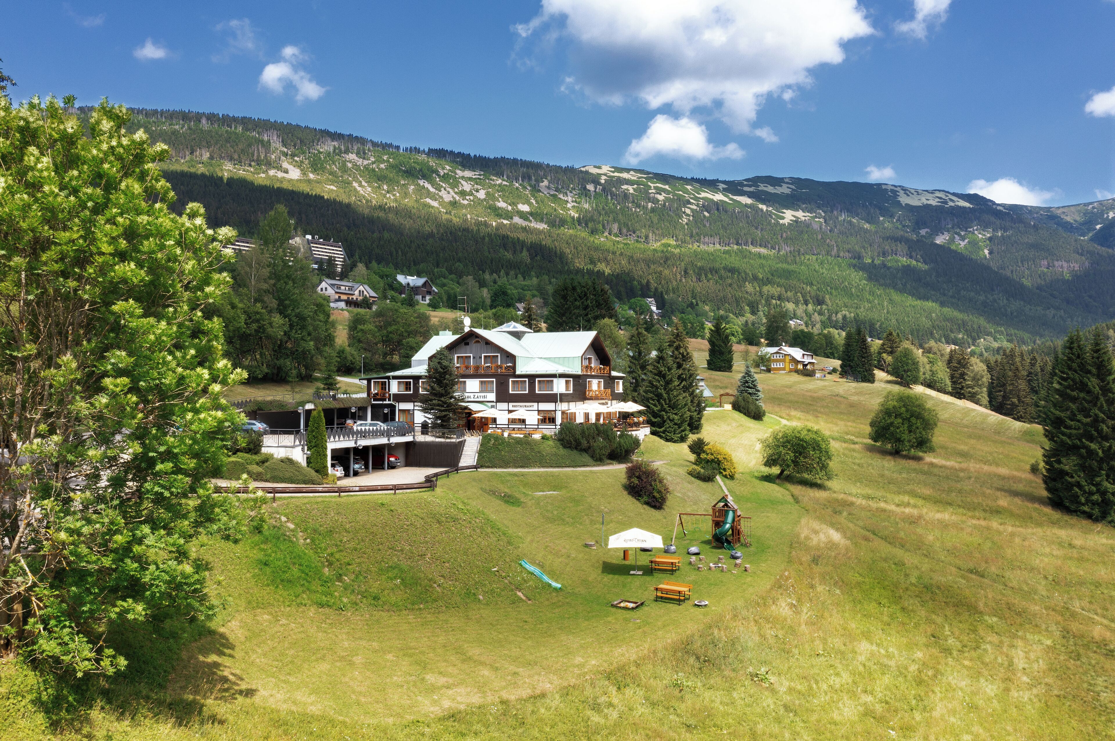 Una casa grande con un amplio espacio exterior, que incluye una terraza y una zona de juegos, rodeada de un paisaje verde exuberante y montañas.