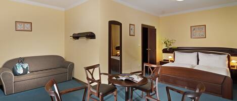 A hotel room with a bed, a sofa, a round table with chairs, and a small table with a vase and a plant.