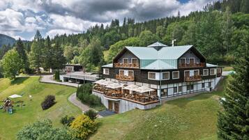 A multi-story hotel with a balcony, stone foundation, and a view of a forested hillside.