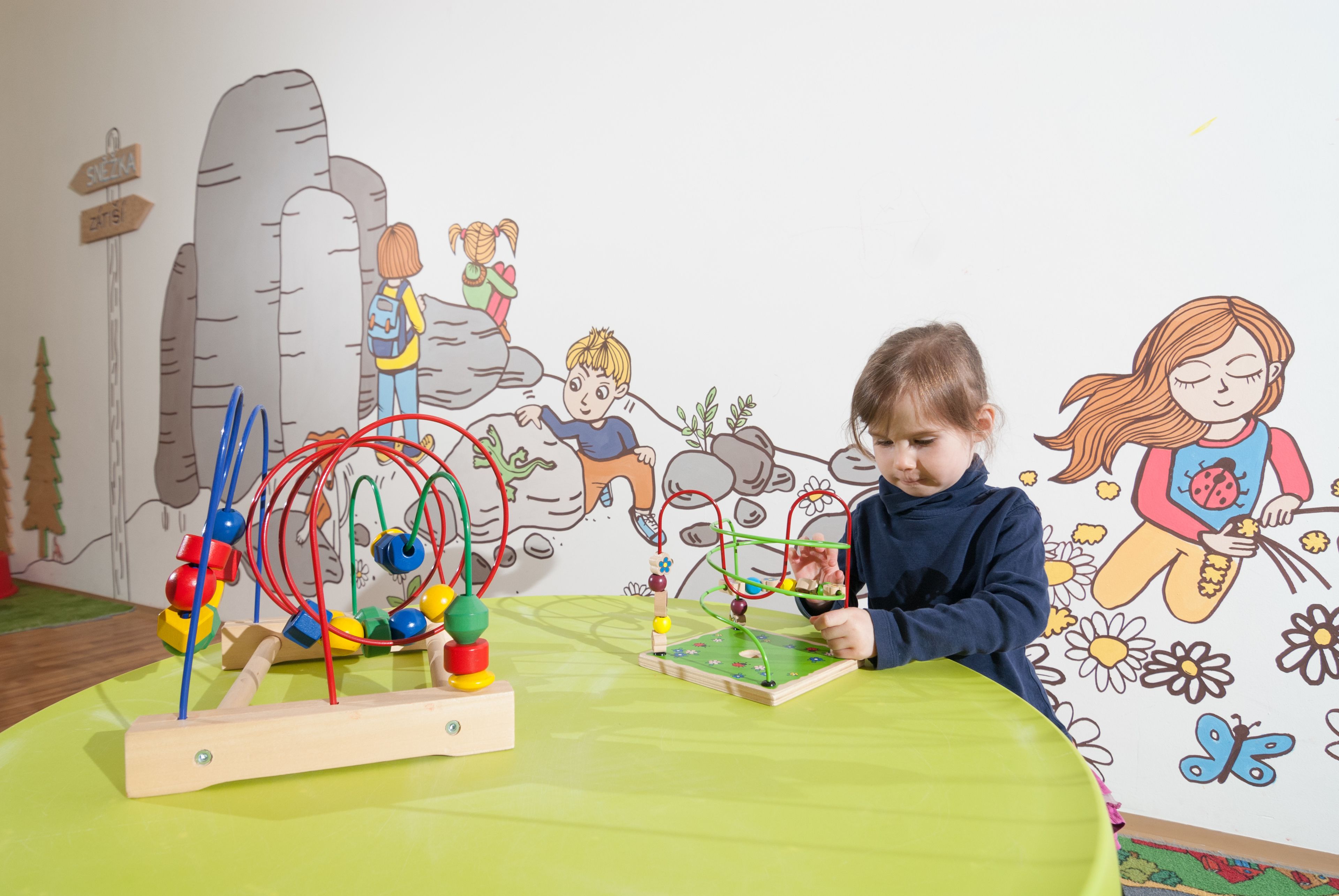 A child playing with a wooden toy on a green table in a room with a wall mural.