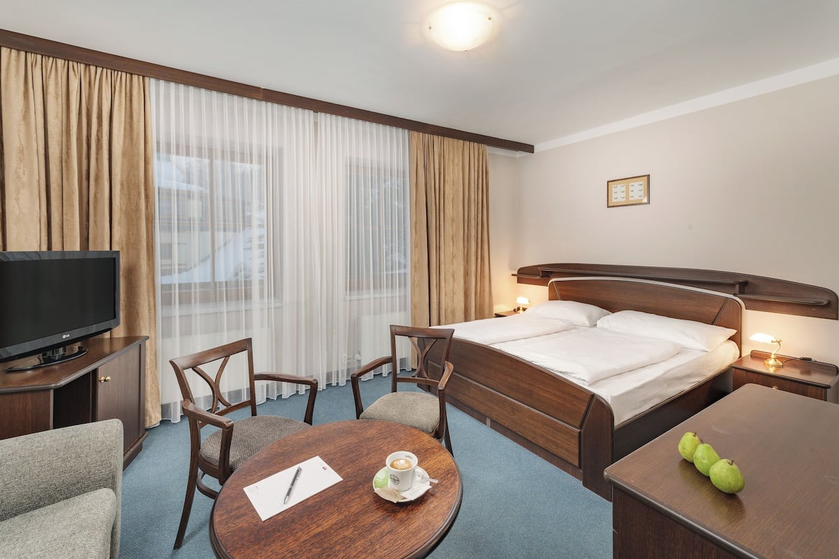 Quarto de hotel com cama, televisão, escrivaninha com cadeira, mesa redonda com uma chávena e caneta, e uma fruta pequena sobre a mesa.