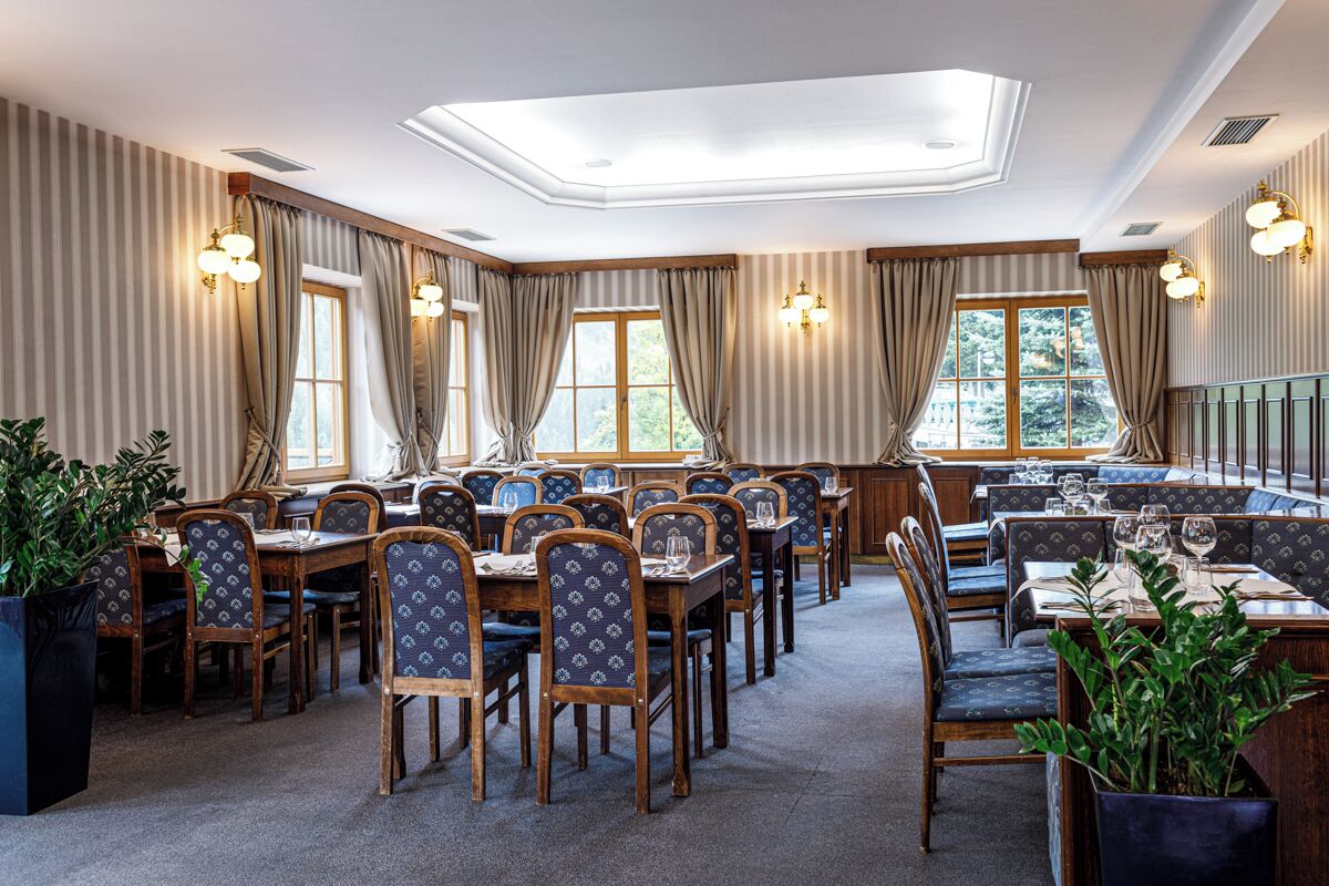 A dining area with multiple tables and chairs, large windows with curtains, and ceiling lights.