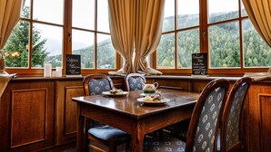 A dining area with a wooden table and chairs, a view of a forest through large windows, and a sign with menu items. - Hotel Zátiší (Spindleruv Mlyn)
