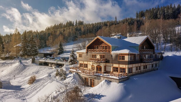 A large, multi-story lodge with a wooden exterior and balconies, surrounded by snow-covered terrain and trees. - Hotel Zátiší (Spindleruv Mlyn)