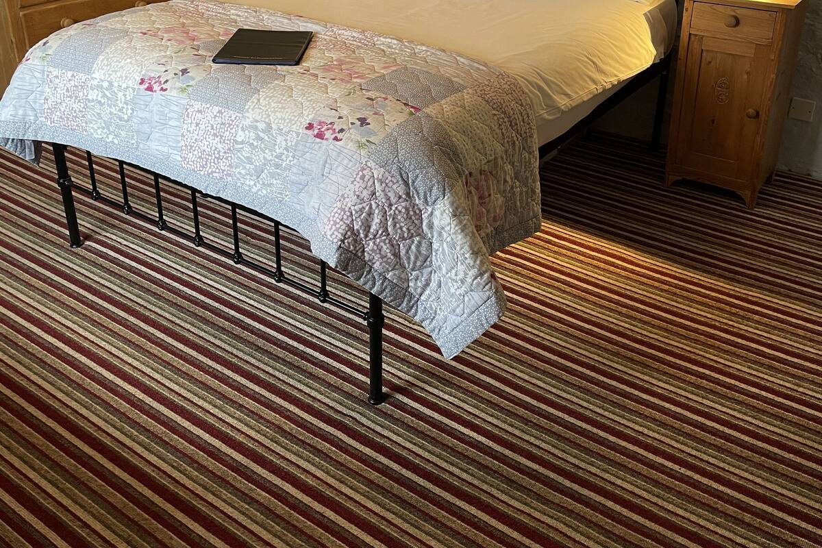 A neatly made bed with a floral bedspread, two bedside tables with lamps, a wooden nightstand, and a striped carpet.