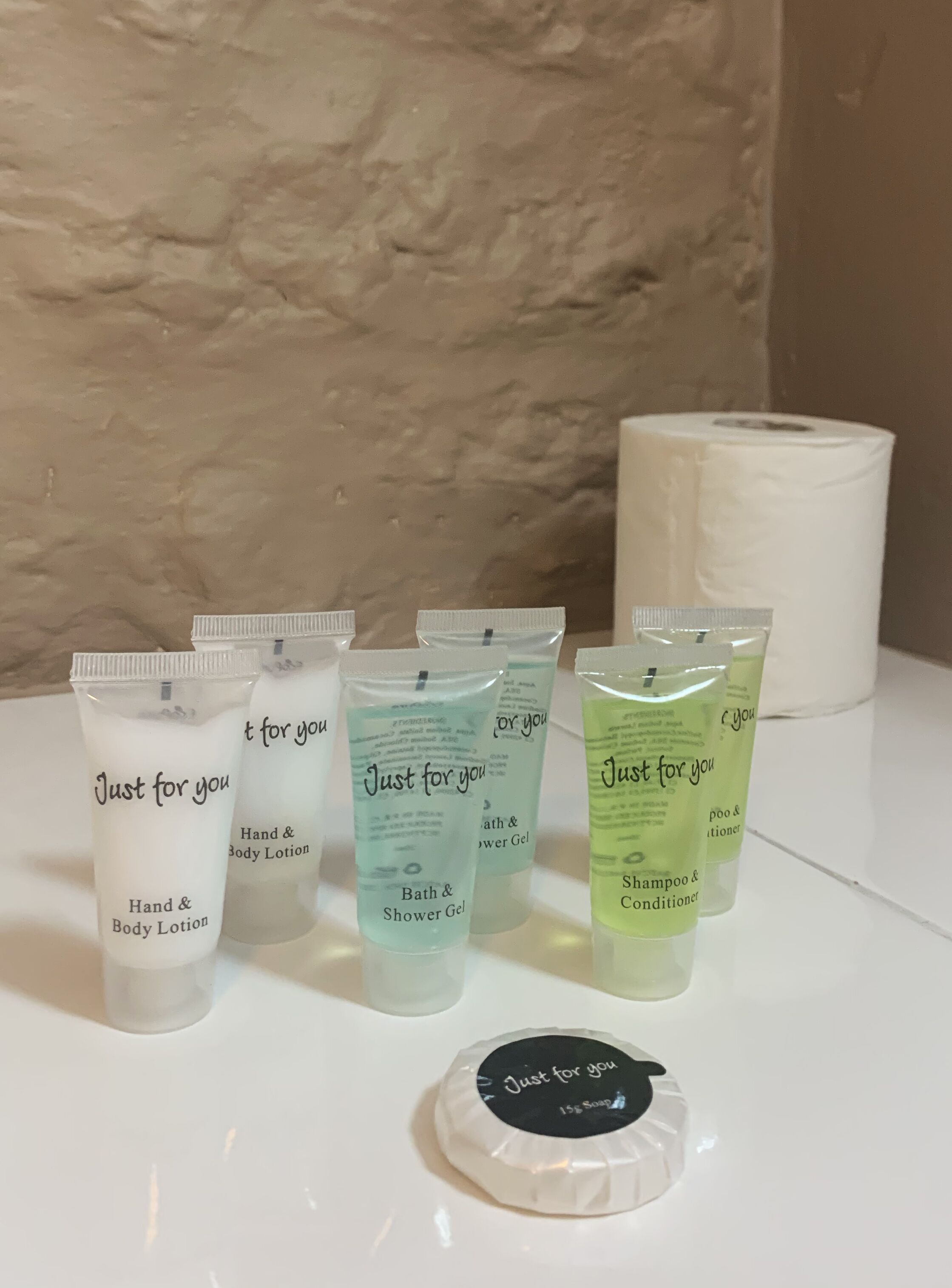 Un conjunto de artículos de aseo que incluye loción corporal y para manos, gel de baño y ducha, champú y acondicionador, y jabón de 15 gramos sobre una superficie blanca, con una pared beige texturizada de fondo.