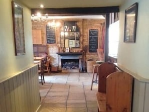 A narrow hallway leading to a dining area with wooden benches and a brick wall. There are framed pictures on the walls and a chalkboard menu visible. - The Royal Oak (Burford)