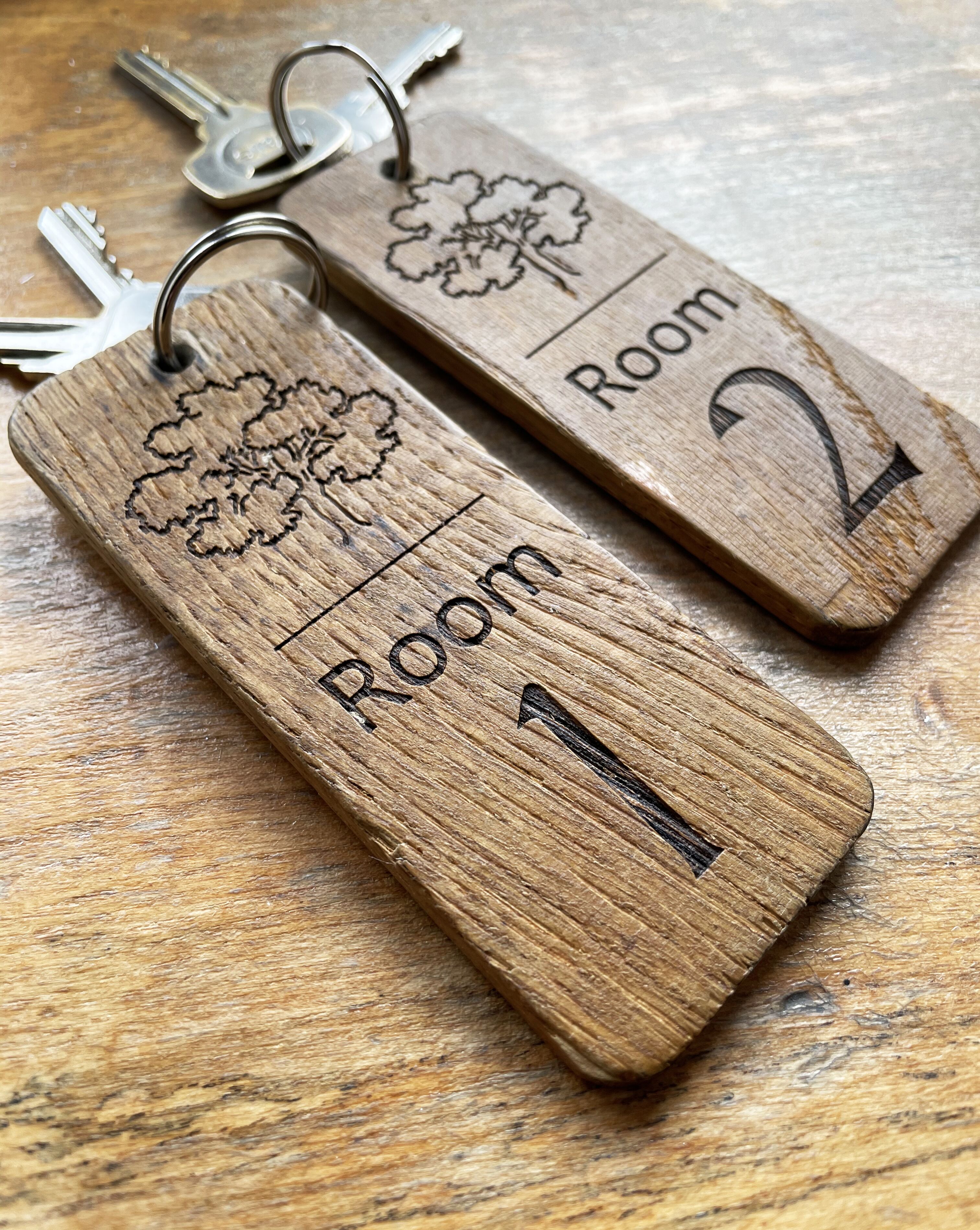 Deux porte-clés en bois, ornés de motifs représentant des arbres et portant les numéros des chambres, sont posés sur une surface en bois.
