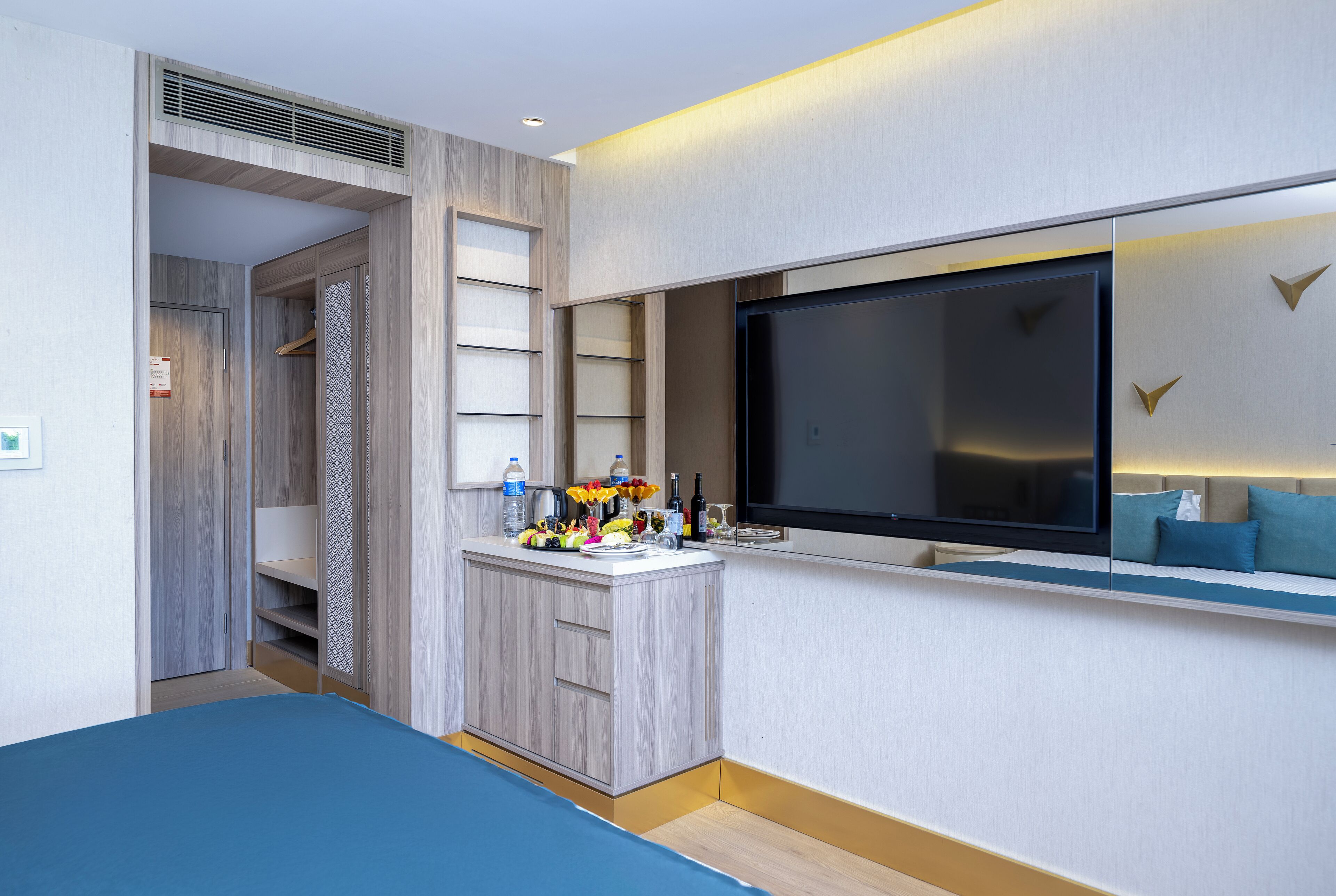 Una habitación de hotel moderna con cama, una kitchenette pequeña, televisión y un área de descanso con sofá y almohadas.