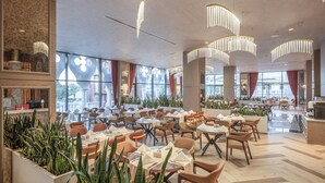 Uma ampla sala de jantar com várias mesas e cadeiras, grandes janelas e luminárias modernas.