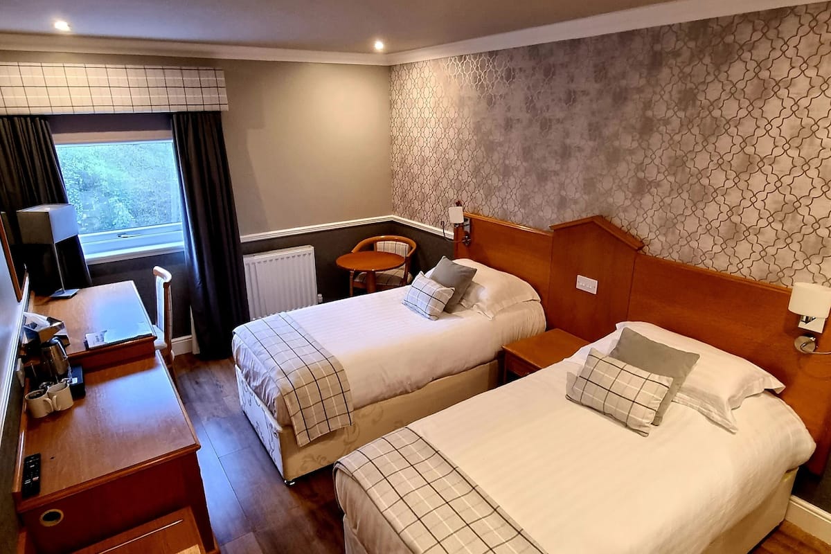 A hotel room with two beds, a desk, a chair, a window with curtains, a radiator, and a patterned wall.