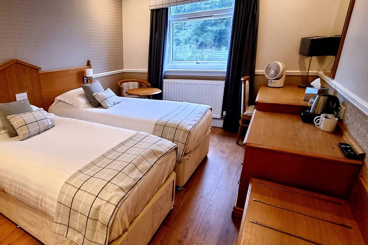 A hotel room with two beds, a wooden nightstand, a small table, a desk with a fan, and a window with a view of trees.