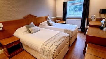 Une chambre d’hôtel avec deux lits, une table de chevet en bois, une petite table, un bureau avec un ventilateur et une fenêtre donnant sur des arbres.