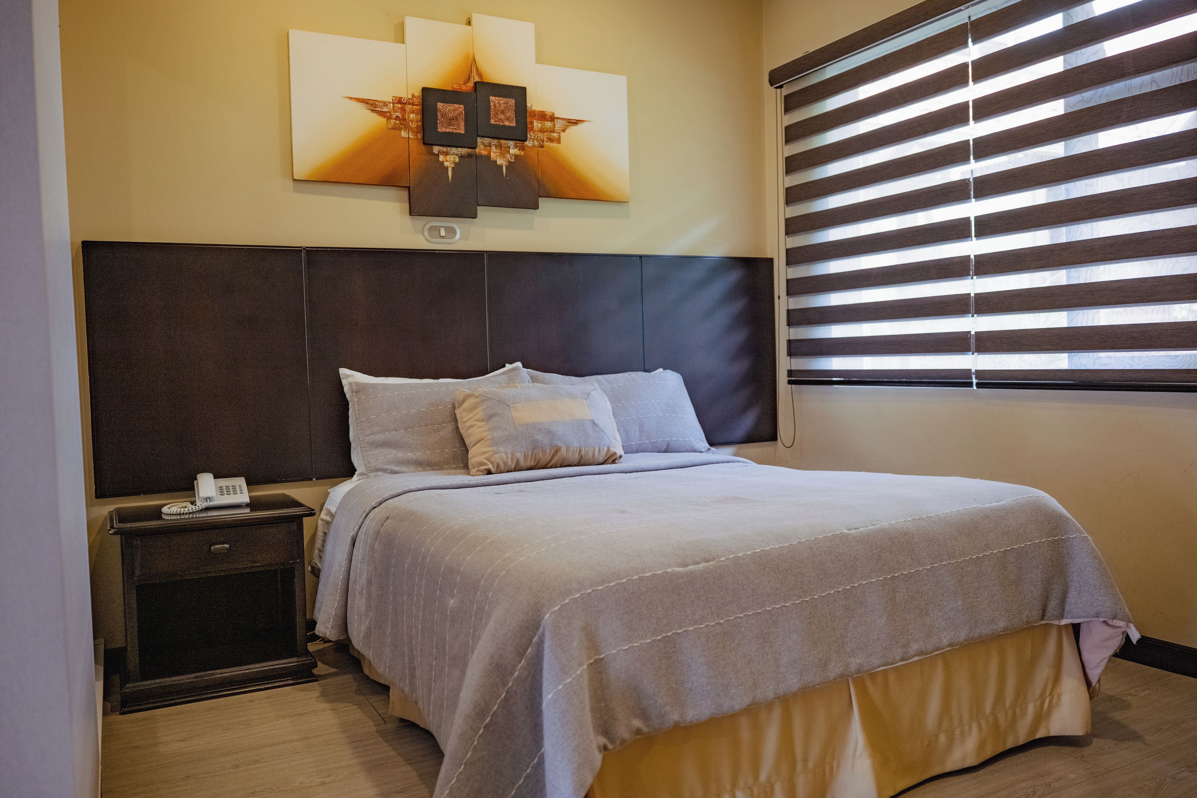 Habitación de hotel con cama, mesita de noche, teléfono en la pared y ventana con persianas.