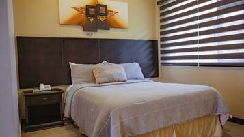 Habitación de hotel con cama, mesita de noche, teléfono en la pared y ventana con persianas.