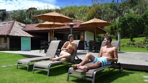 Two people relaxing on sun loungers with umbrellas, a stone building, and a garden in the background. - Hosteria Durán (Banos)