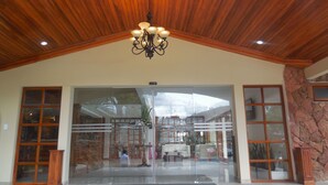 A hotel lobby with a wooden ceiling, a chandelier, large glass doors, and a stone wall. - Hosteria Durán (Banos)