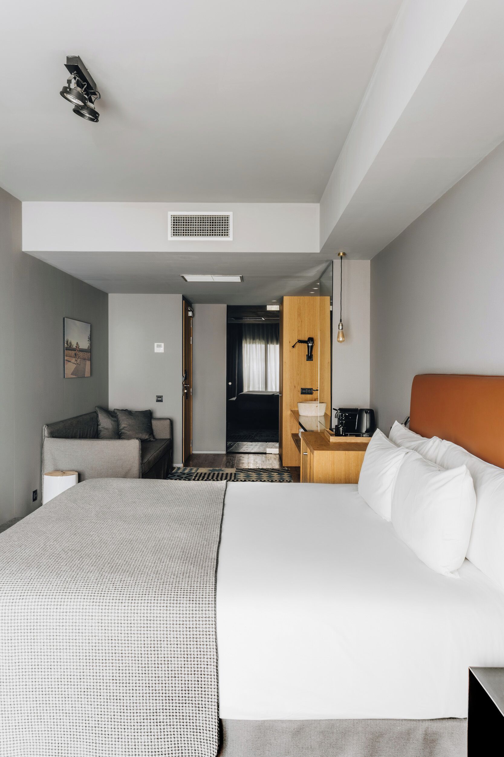 A hotel room with a large bed, a desk, a chair, a sofa, a coffee machine, a mirror, and a door leading to another room.