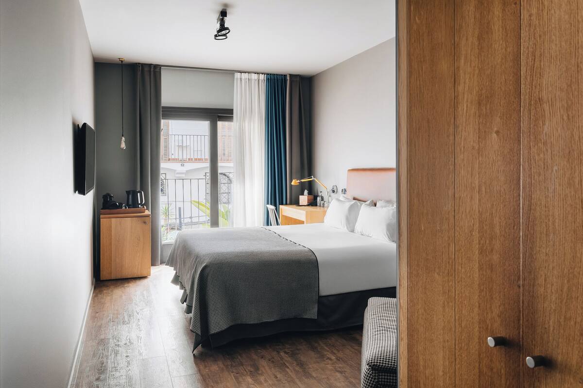 Een hotelkamer met een bed, nachtkastje, lamp en uitzicht op het exterieur.