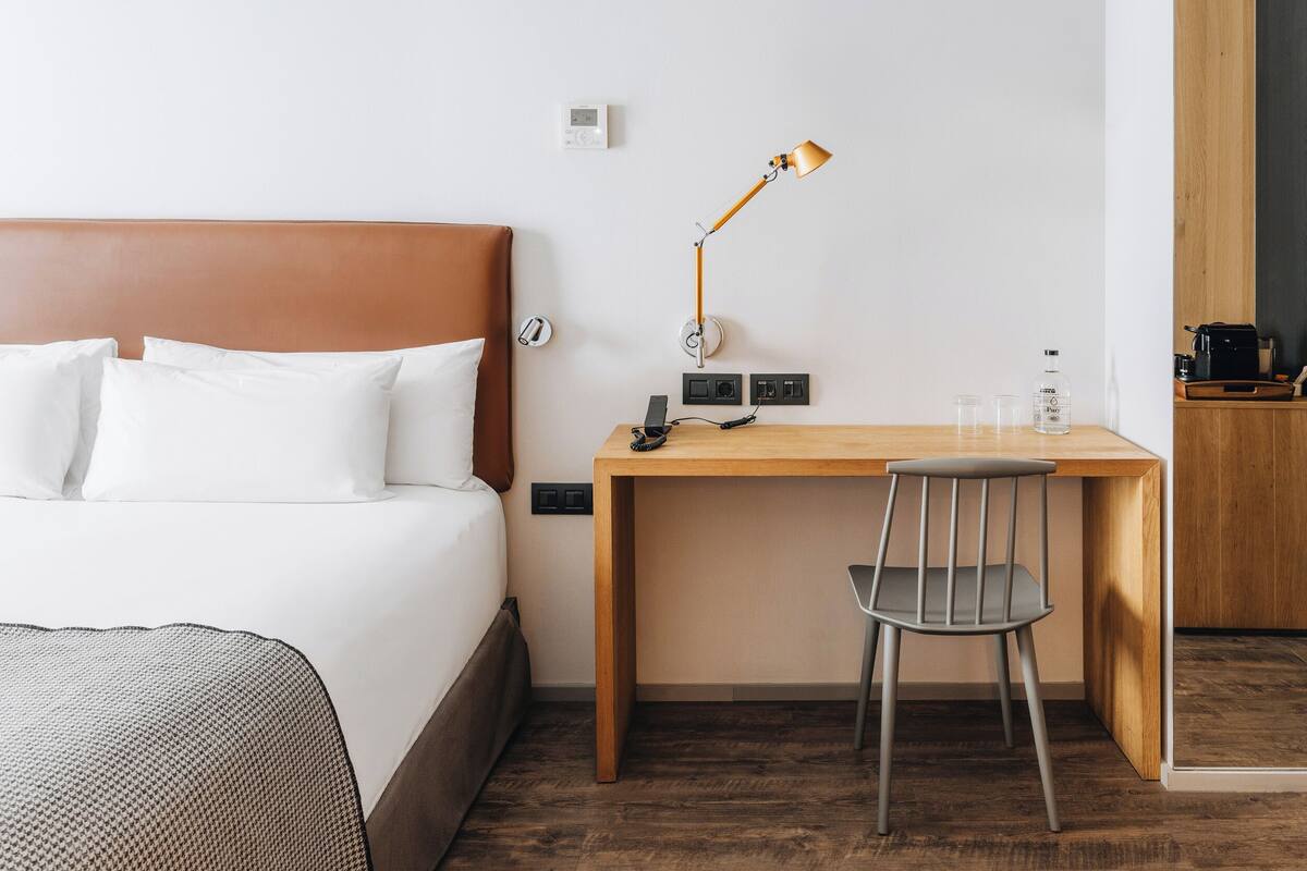 Een hotelkamer met een bed, een bureau, een stoel, een lamp, een thermostaat en een koffiezetapparaat.