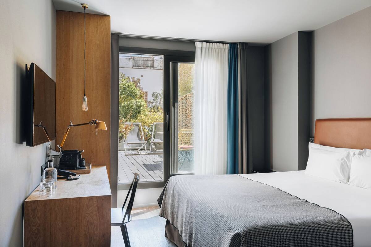Hotellihuone, jossa on sänky, työpöytä lampun kanssa, televisio ja näkymä ulos.