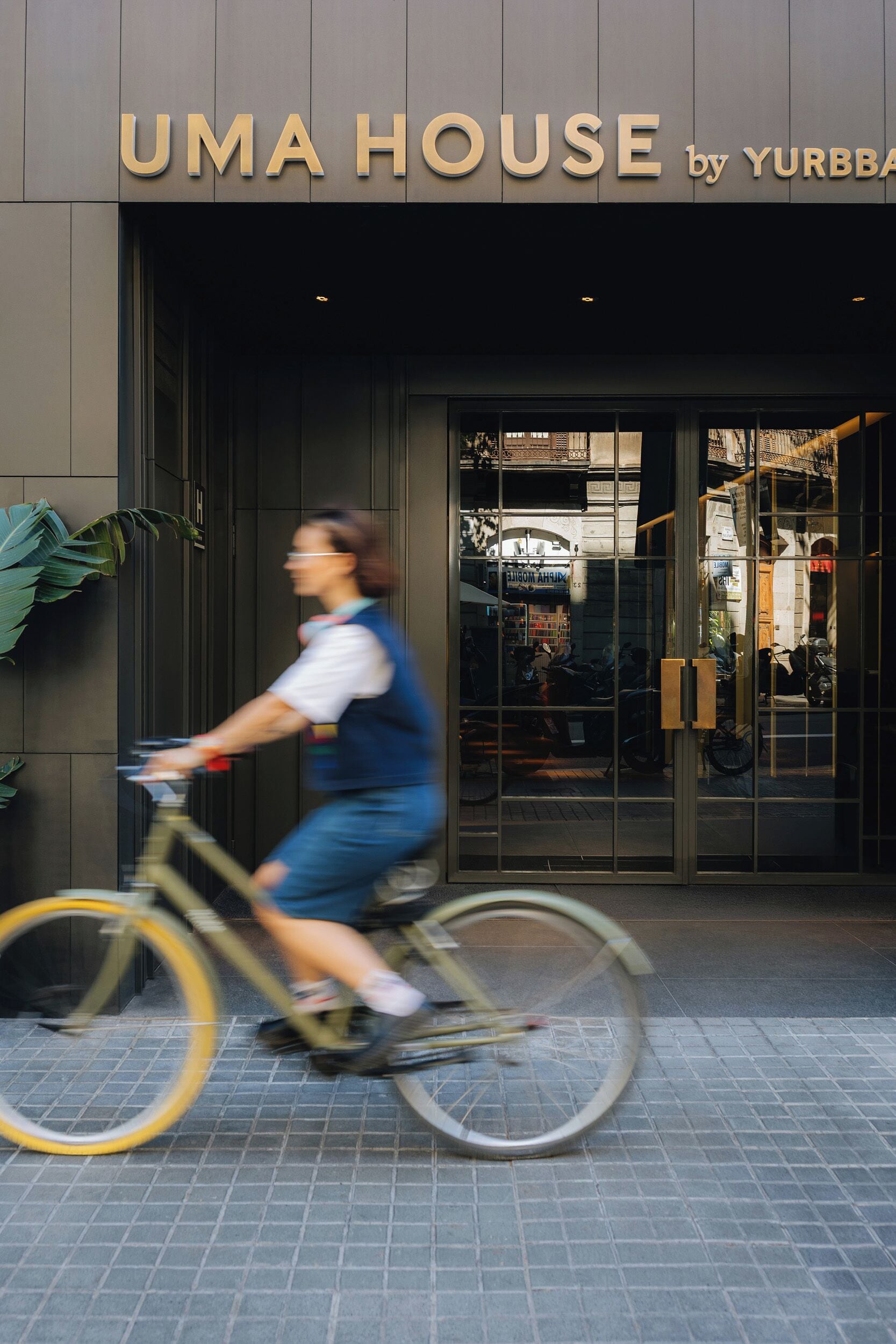 Een winkelgevel met het bord 'UMA HOUSE by YURBBA' en een persoon die op een fiets voorbijrijdt.