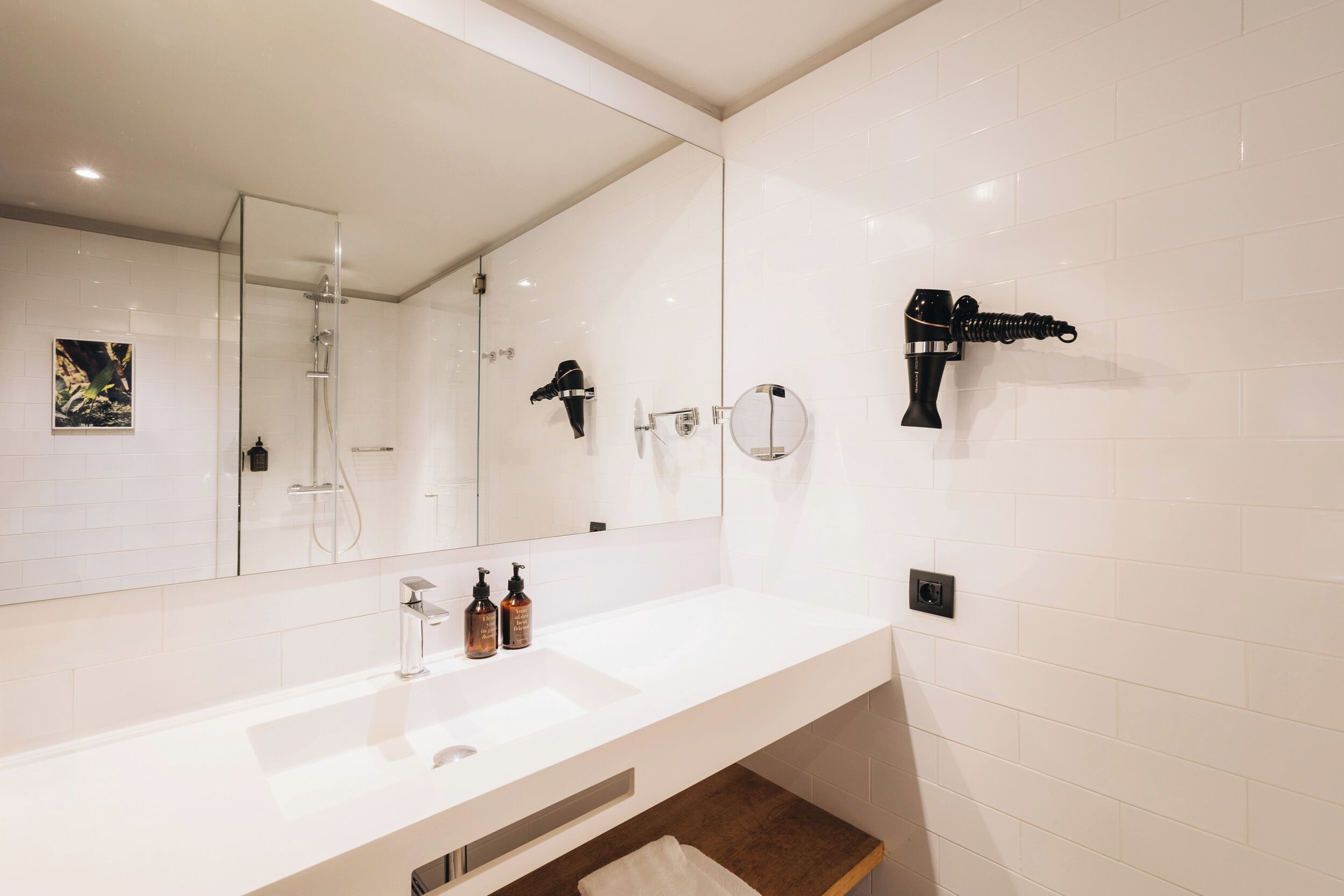 Un baño moderno con un lavamanos blanco, un espejo grande, un secador de pelo portátil y una ducha con mampara de vidrio.