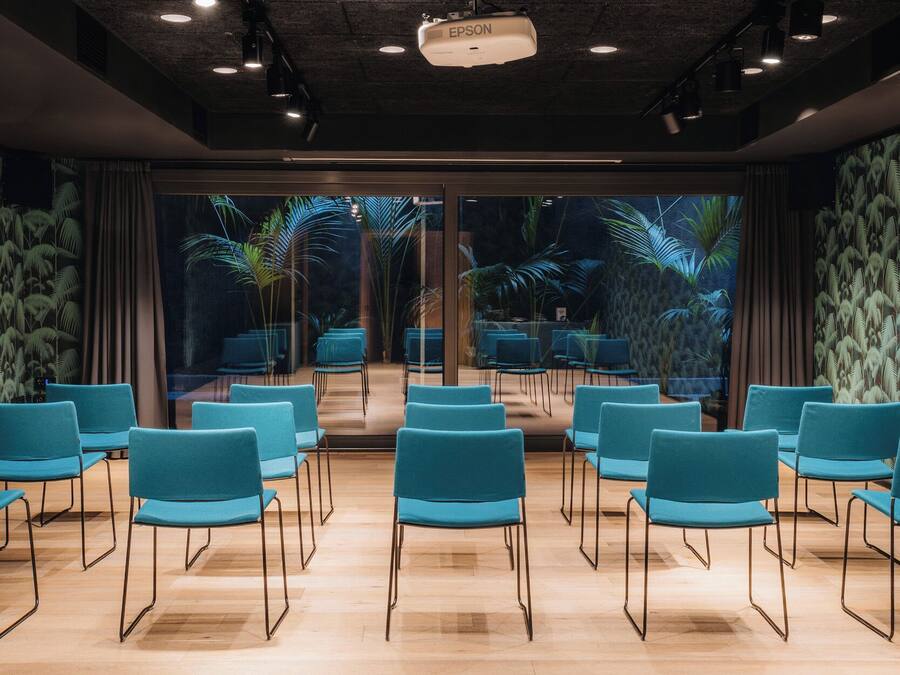 A conference room with rows of chairs, a projector, and a view of tropical plants through large windows.