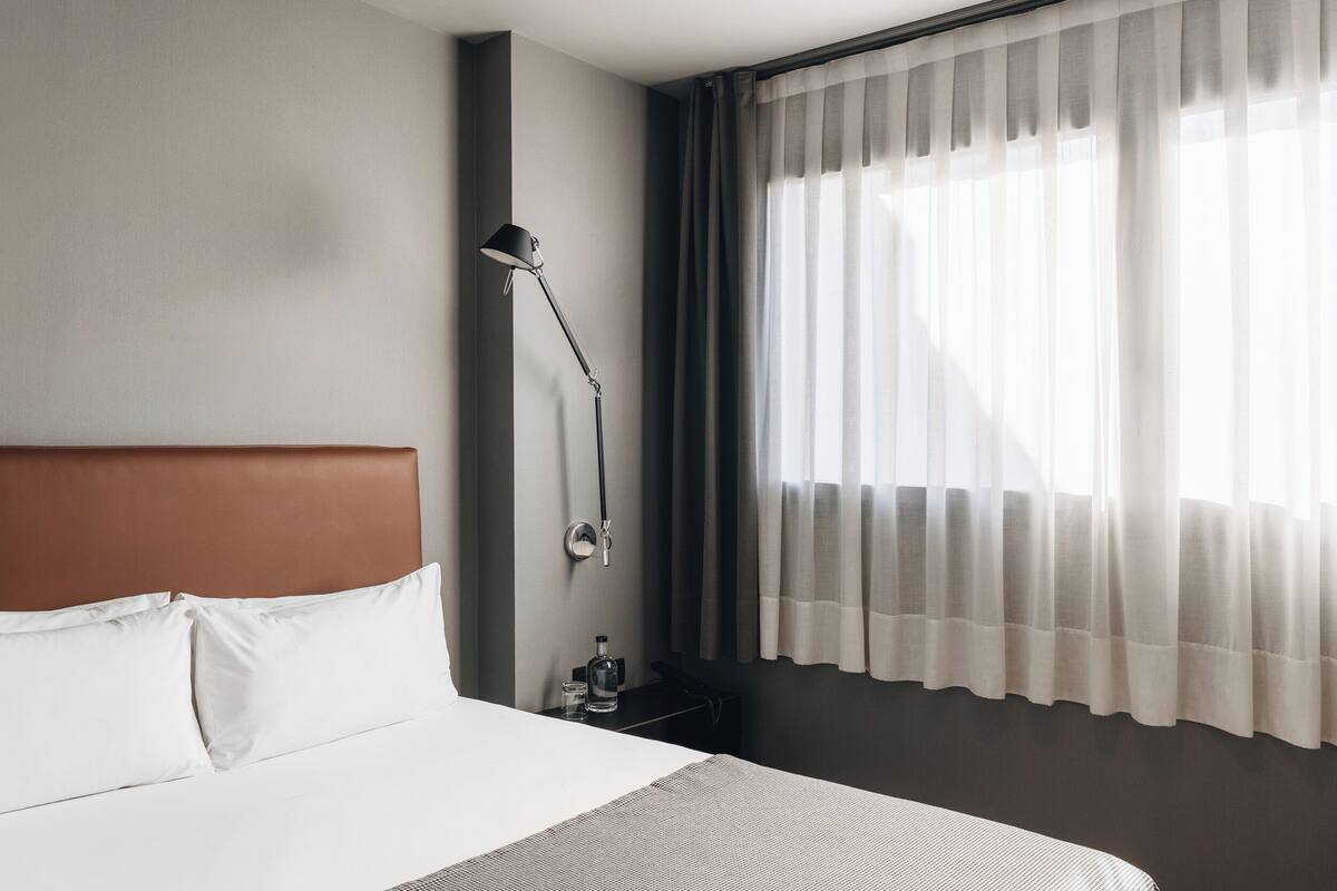 Een hotelkamer met een bed, een nachtkastje met een lamp en een raam met gordijnen.