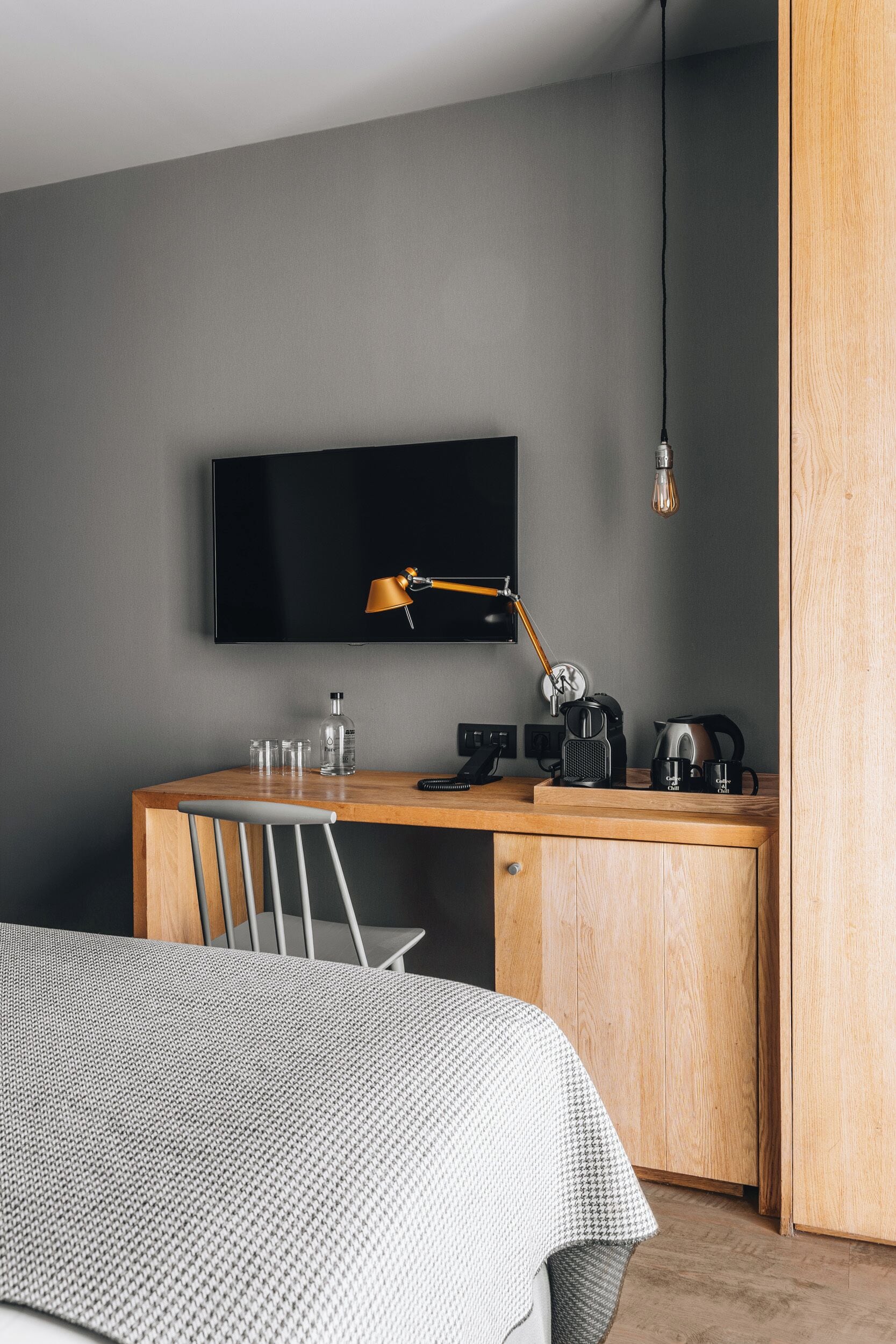 A modern room with a wooden desk, a mounted TV, a lamp, and a bed with a patterned blanket.