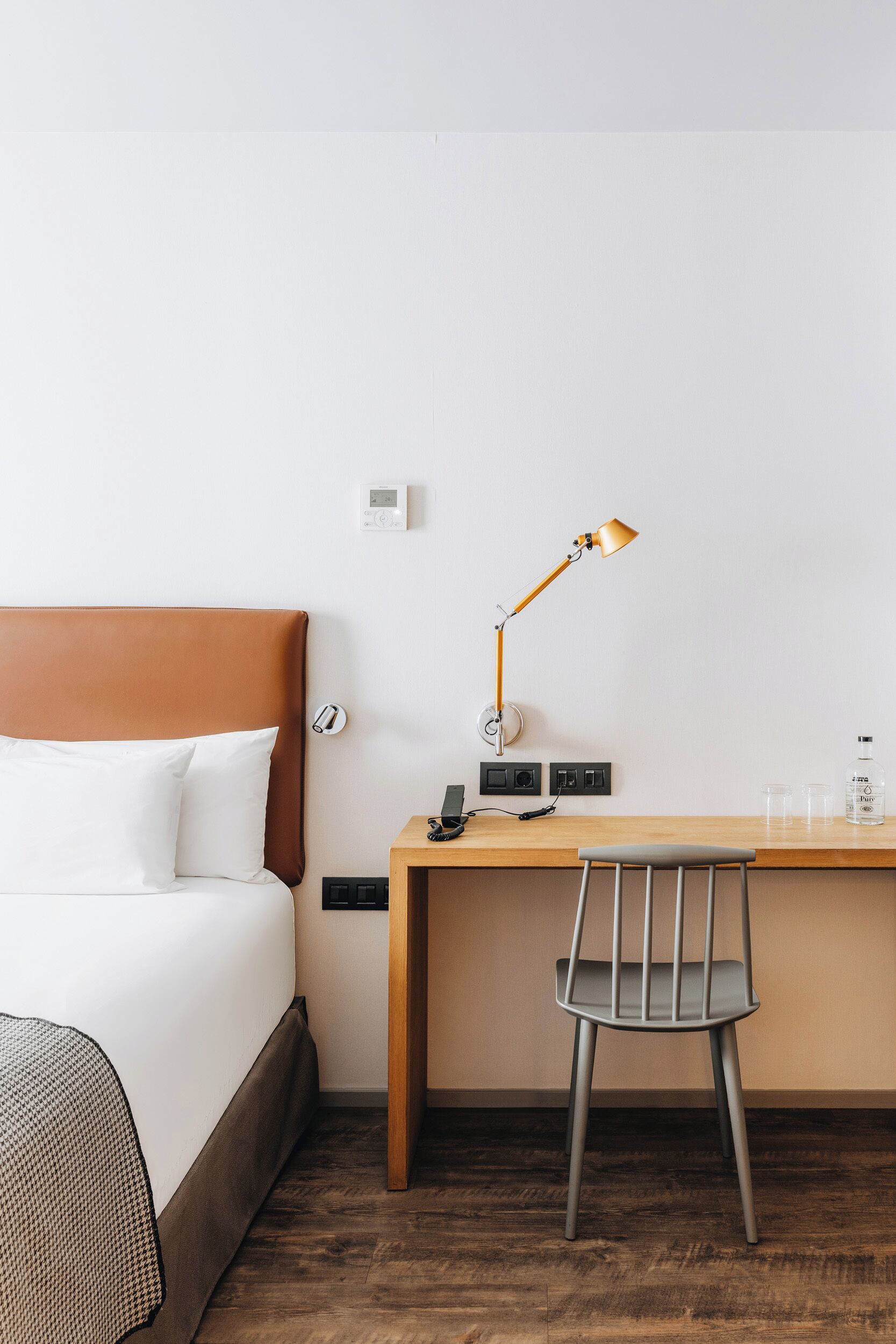 A hotel room with a bed, a desk, a chair, a lamp, a thermostat, and a water bottle.