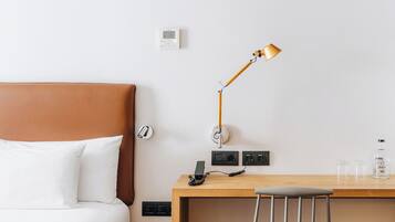 A hotel room with a bed, a desk, a chair, a lamp, a thermostat, and a water bottle.