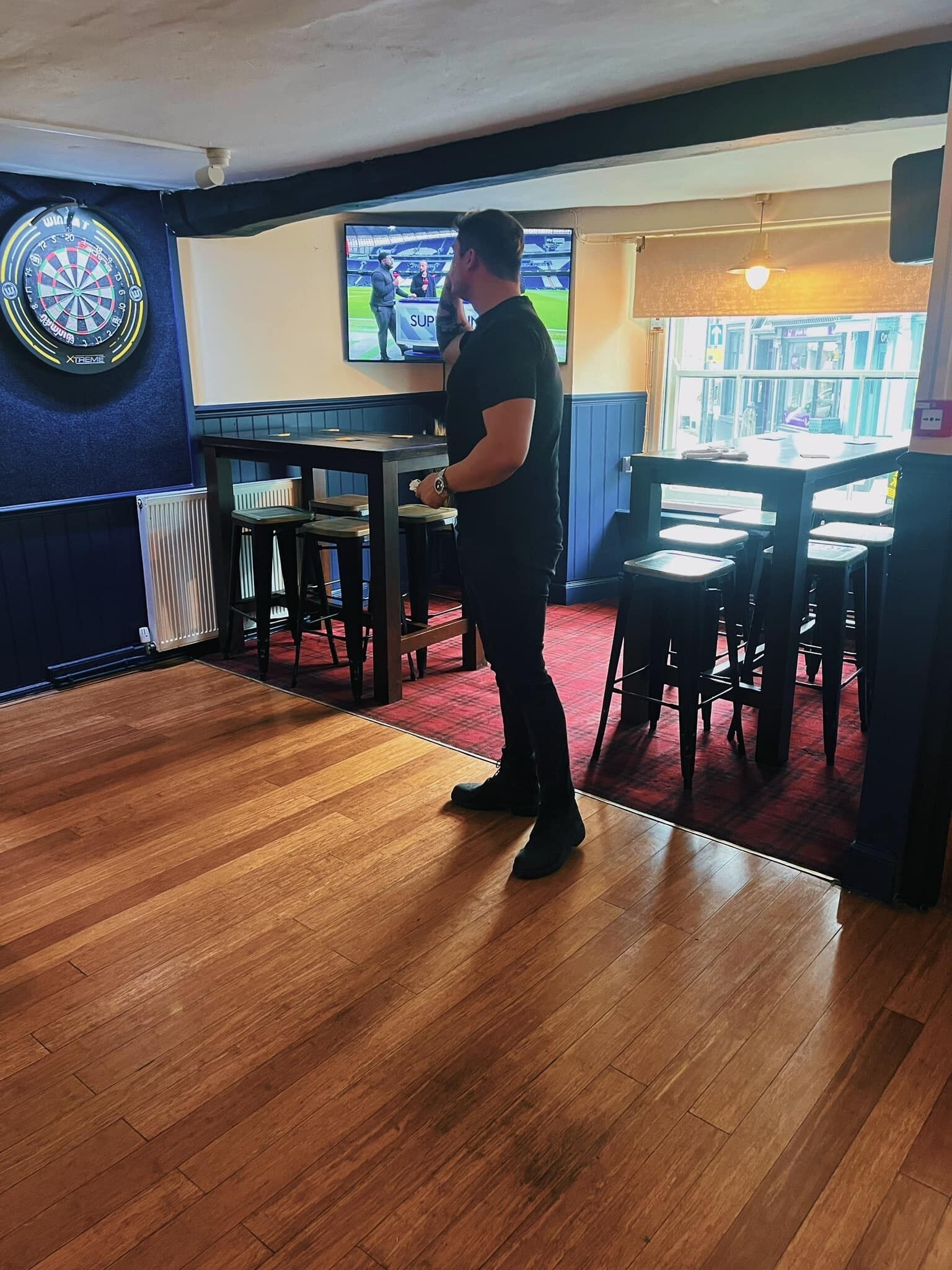 A man standing in a room with a dartboard, a television displaying a sports event, and a bar area with stools and a table.