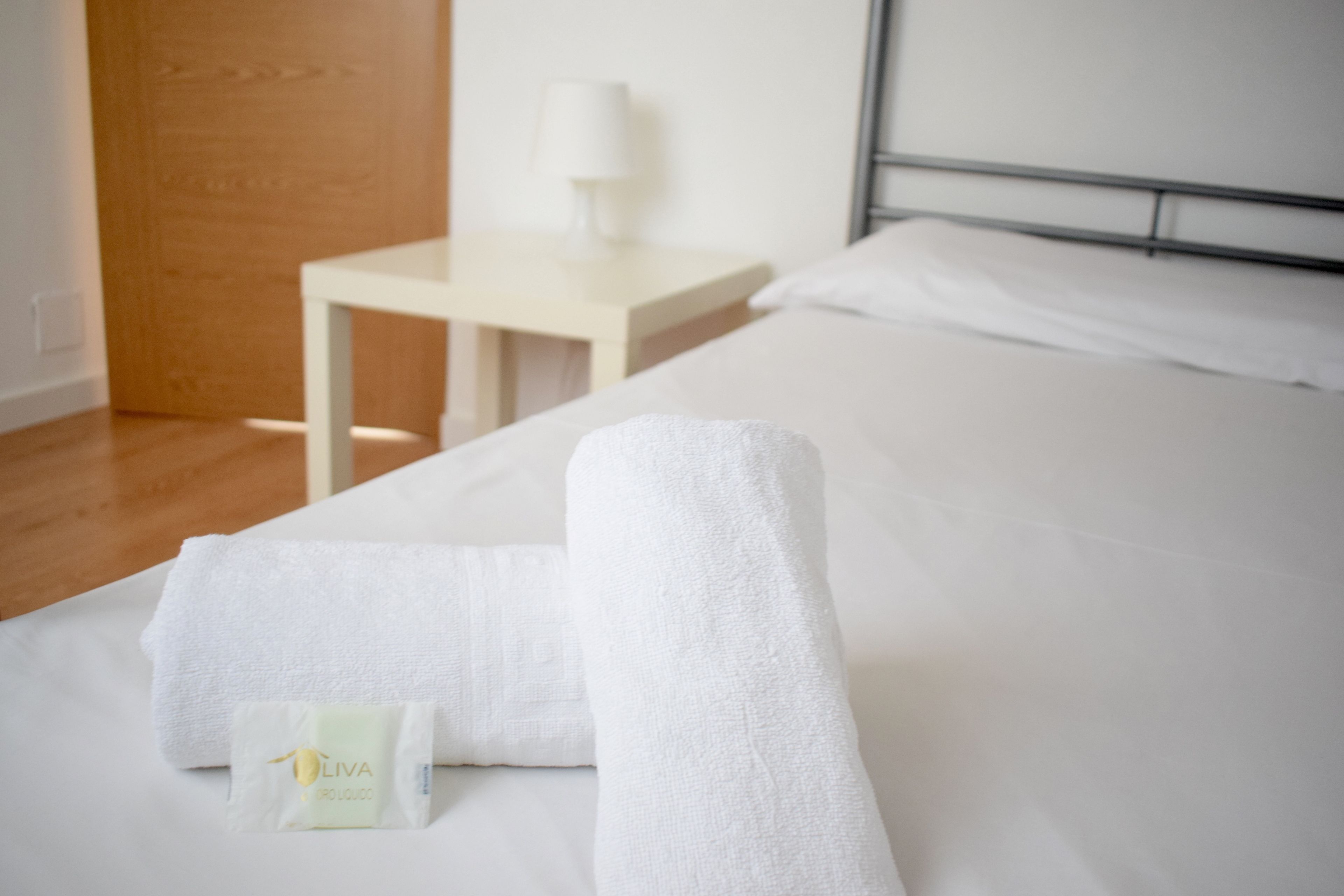 Una cama bien tendida con sábanas blancas y una toalla doblada. Un pequeño paquete con la etiqueta 'OLIVA' está colocado sobre la cama.