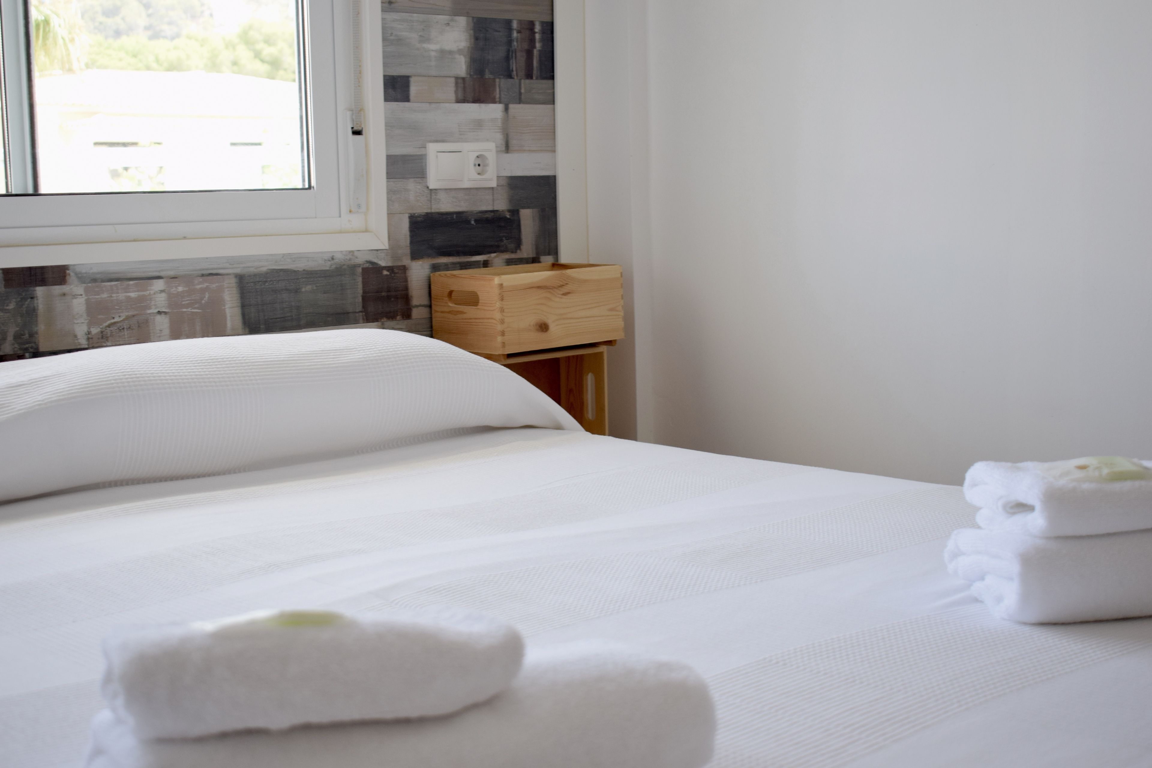 A neatly made bed with white linens and towels, a wooden nightstand with a drawer, and a window with a view of greenery.