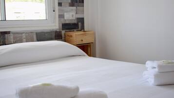 A neatly made bed with white linens and towels, a wooden nightstand with a drawer, and a window with a view of greenery.