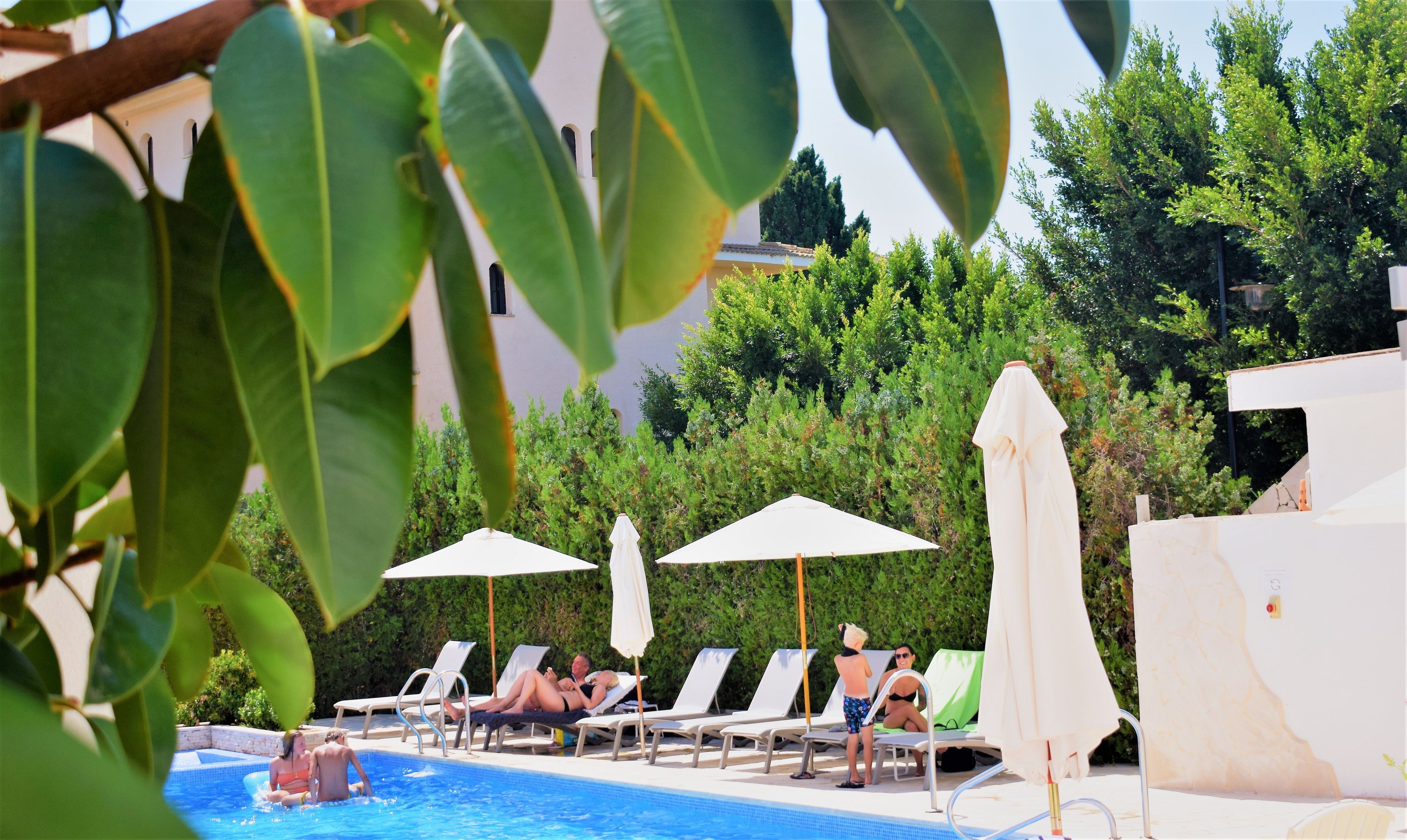 Un espace piscine avec des chaises longues et des parasols, entouré de verdure et avec un bâtiment en arrière-plan.