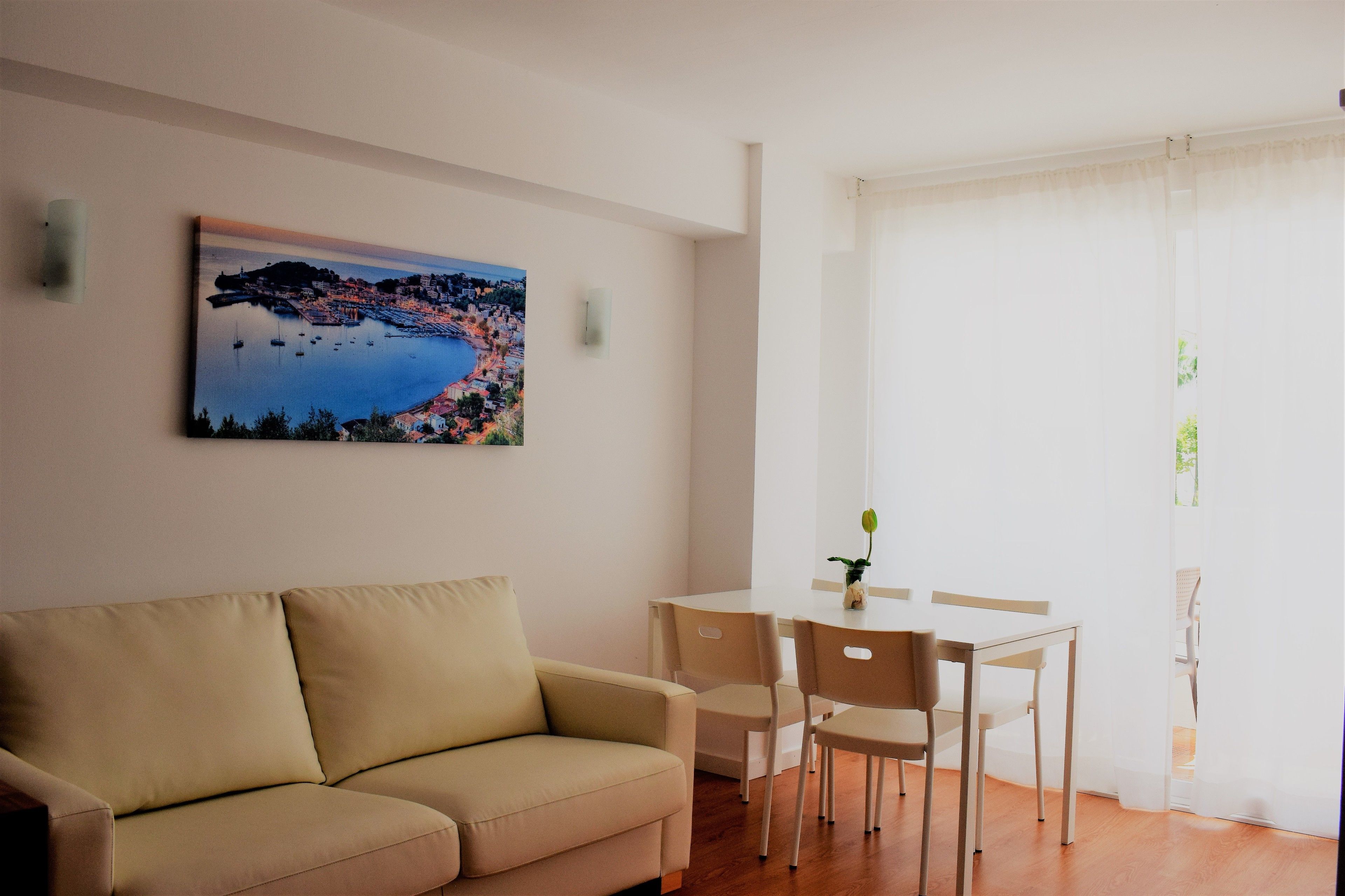 A living room with a beige sofa, a dining table with chairs, and a painting of a coastal town on the wall.