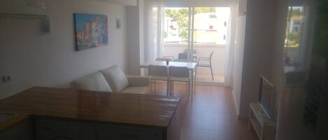 En kompakt opholdsstue med tekøkken, spiseplads med bord og stole, samt udsigt ud af en glasdør.