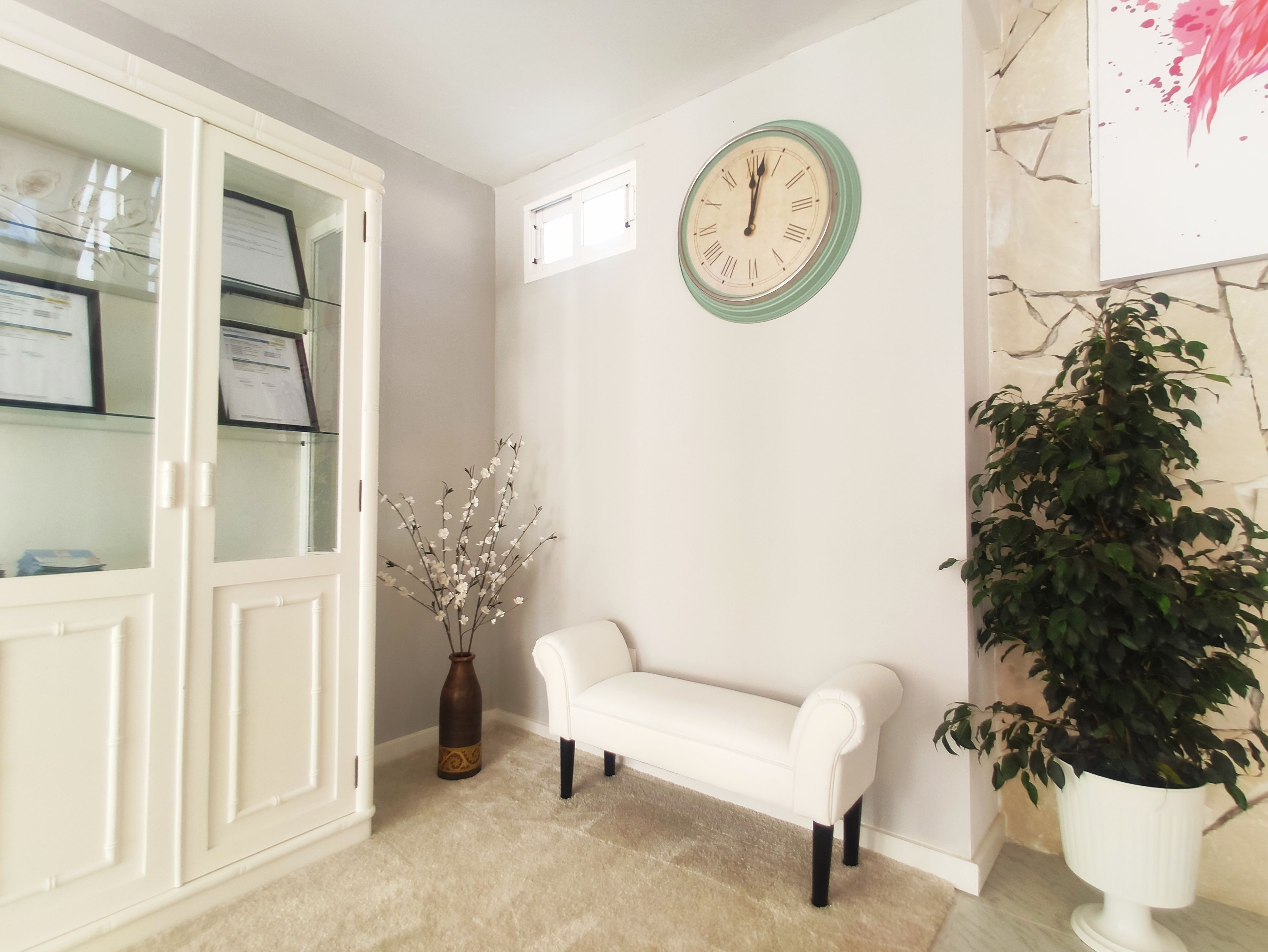 Ein Zimmer mit einer weißen Bank, einer Wanduhr, einer Vase mit Blumen und einem Schrank mit Glastüren.