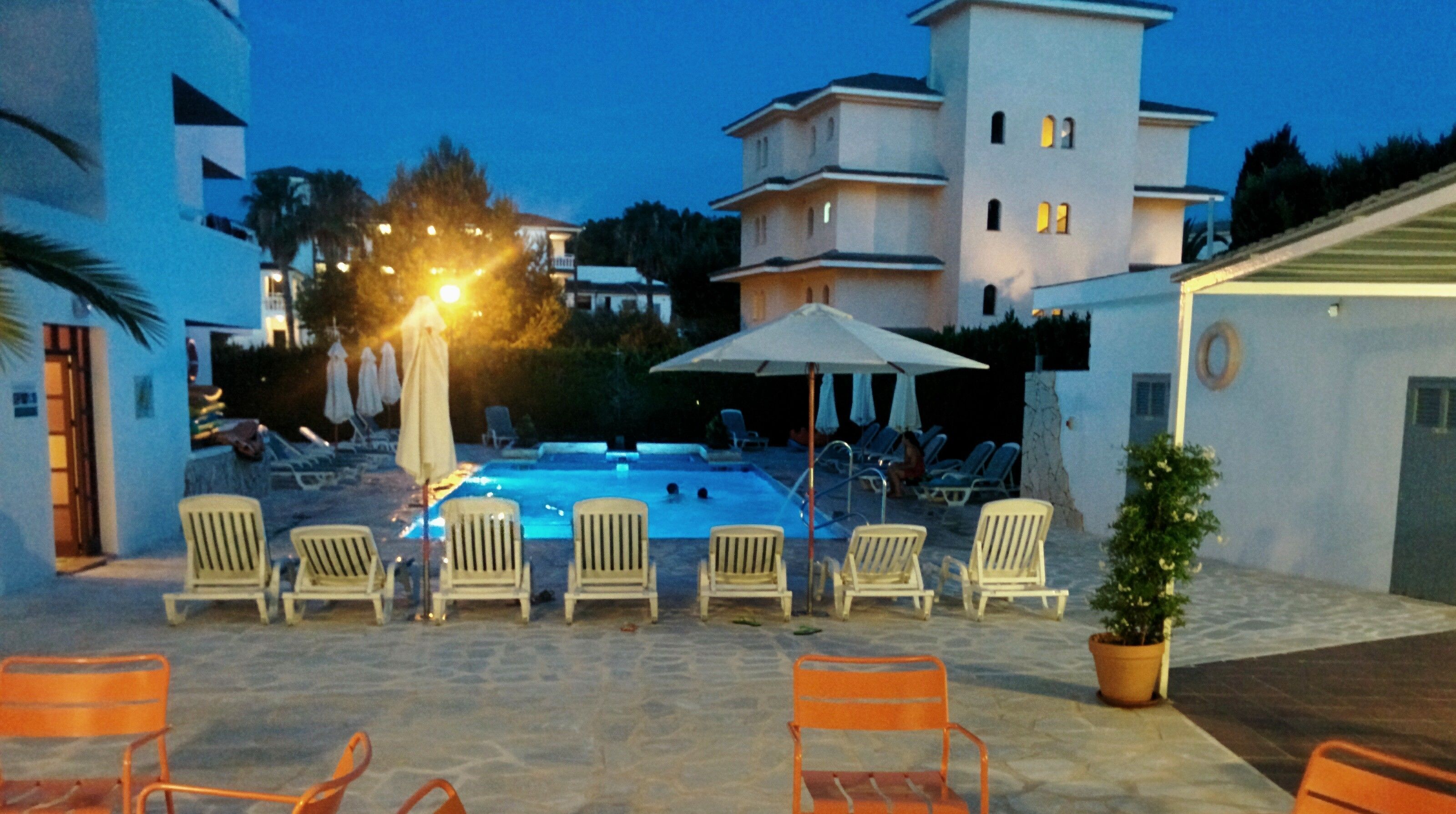 A swimming pool with lounge chairs and umbrellas, surrounded by buildings and a potted plant.