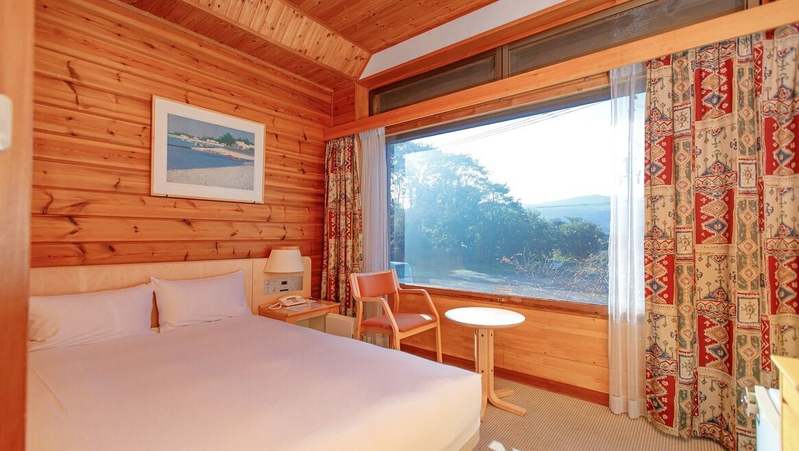 Habitación de cabaña de madera con cama, una mesita, una silla, mesita de noche, lámpara, un cuadro enmarcado y un ventanal con cortinas.