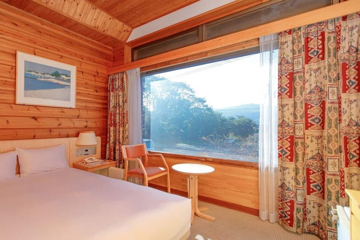 Quarto de cabana de madeira com cama, uma mesinha, uma cadeira, mesa de cabeceira, abajur, quadro emoldurado e uma janela grande com cortinas.