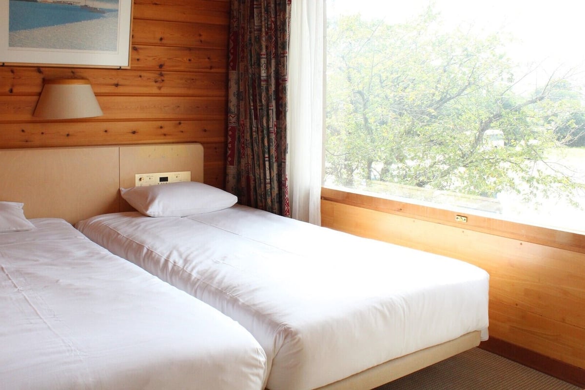 Quarto com duas camas, uma luminária, um quadro e uma janela com vista para árvores.