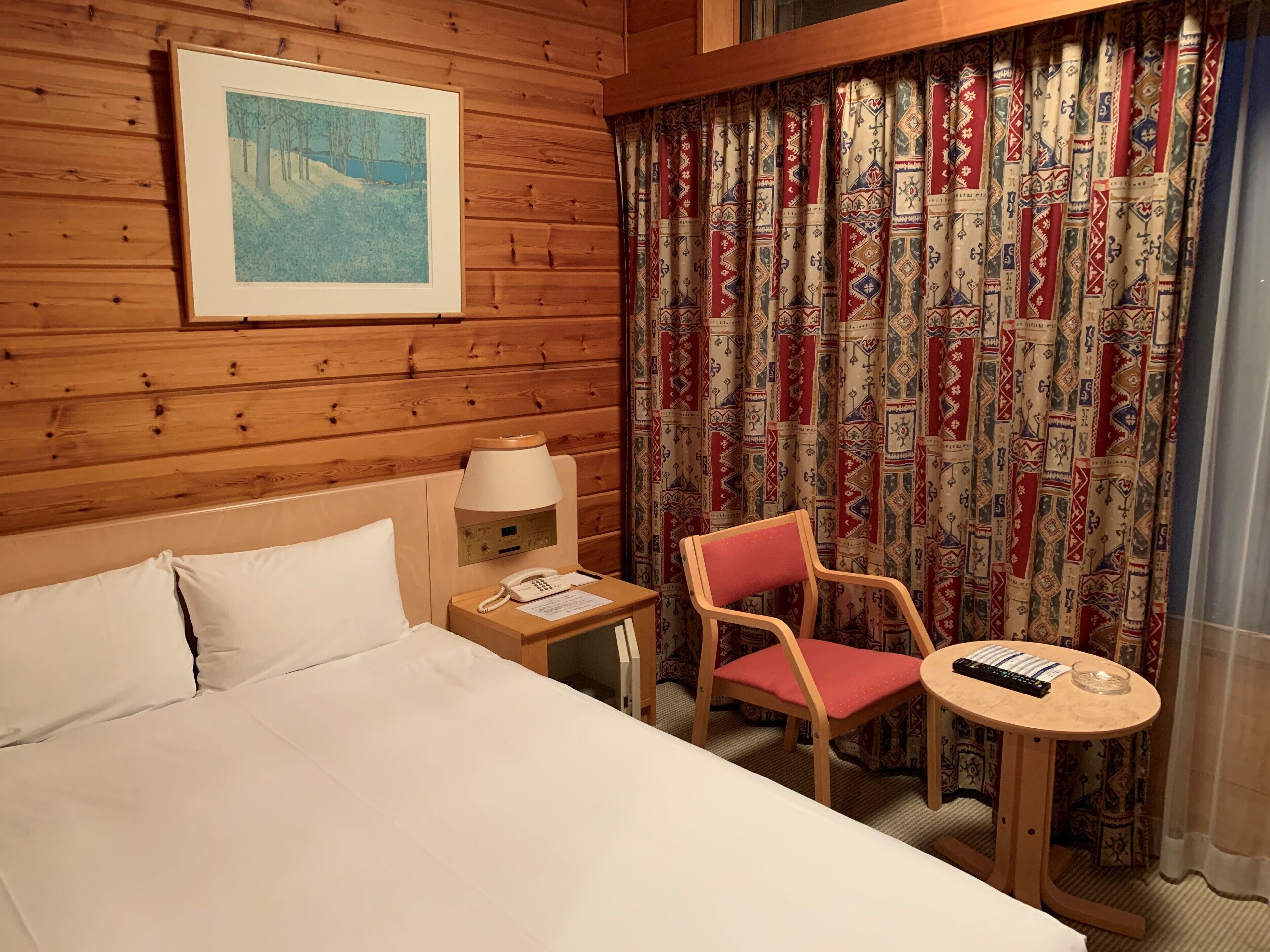 Et hotelværelse med sengegavl i træ, seng med hvide sengetøj, et natbord med lampe og telefon, en rød stol og et lille bord med fjernbetjening.