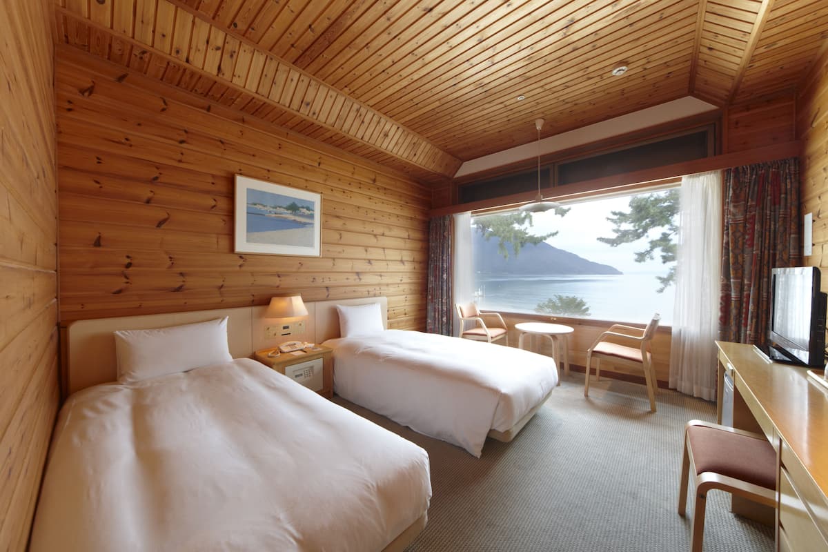 Quarto de cabana de madeira com duas camas, uma escrivaninha, uma cadeira e janela com vista para a água.