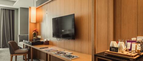 Habitación de hotel con pared de paneles de madera, un televisor de pantalla plana sobre un escritorio de madera, una silla, un atril para revistas y una zona de café.