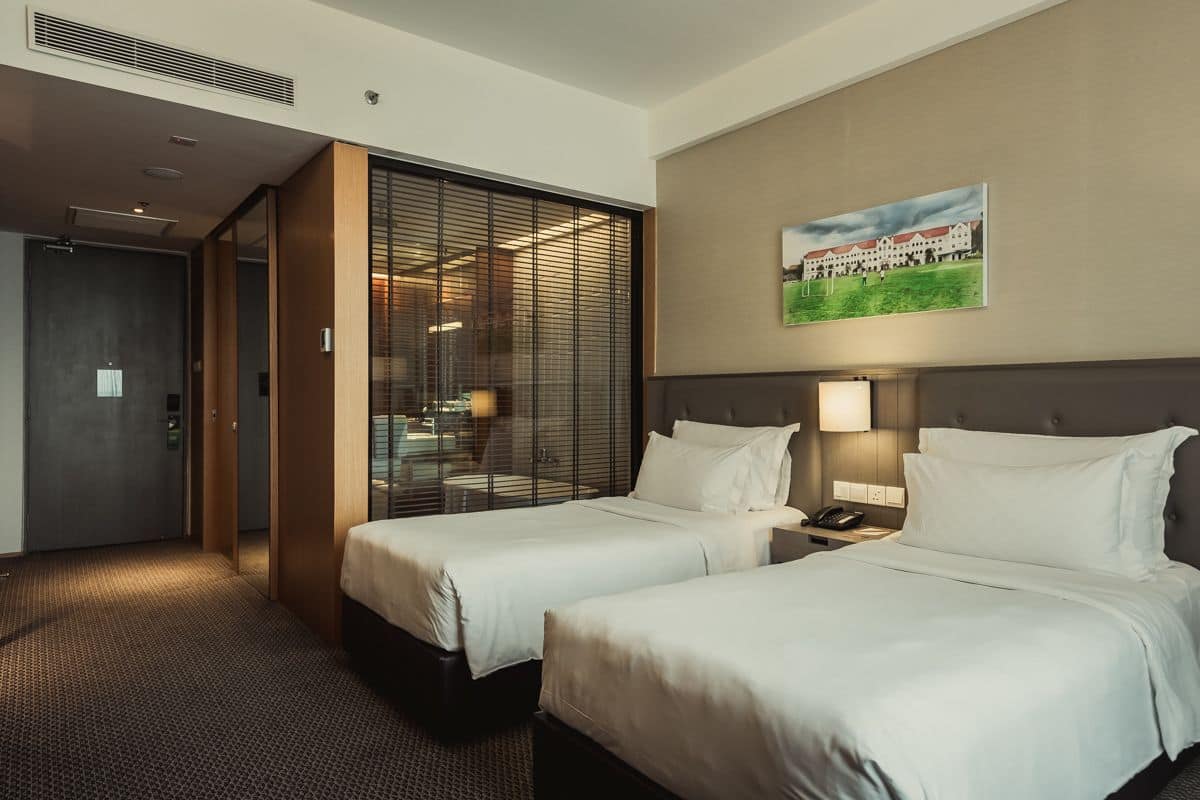 A hotel room with two beds, a large window with blinds, a painting on the wall, and a bedside table with a lamp.