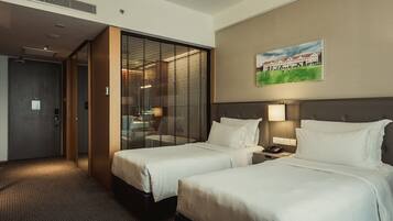 A hotel room with two beds, a large window with blinds, a painting on the wall, and a bedside table with a lamp.