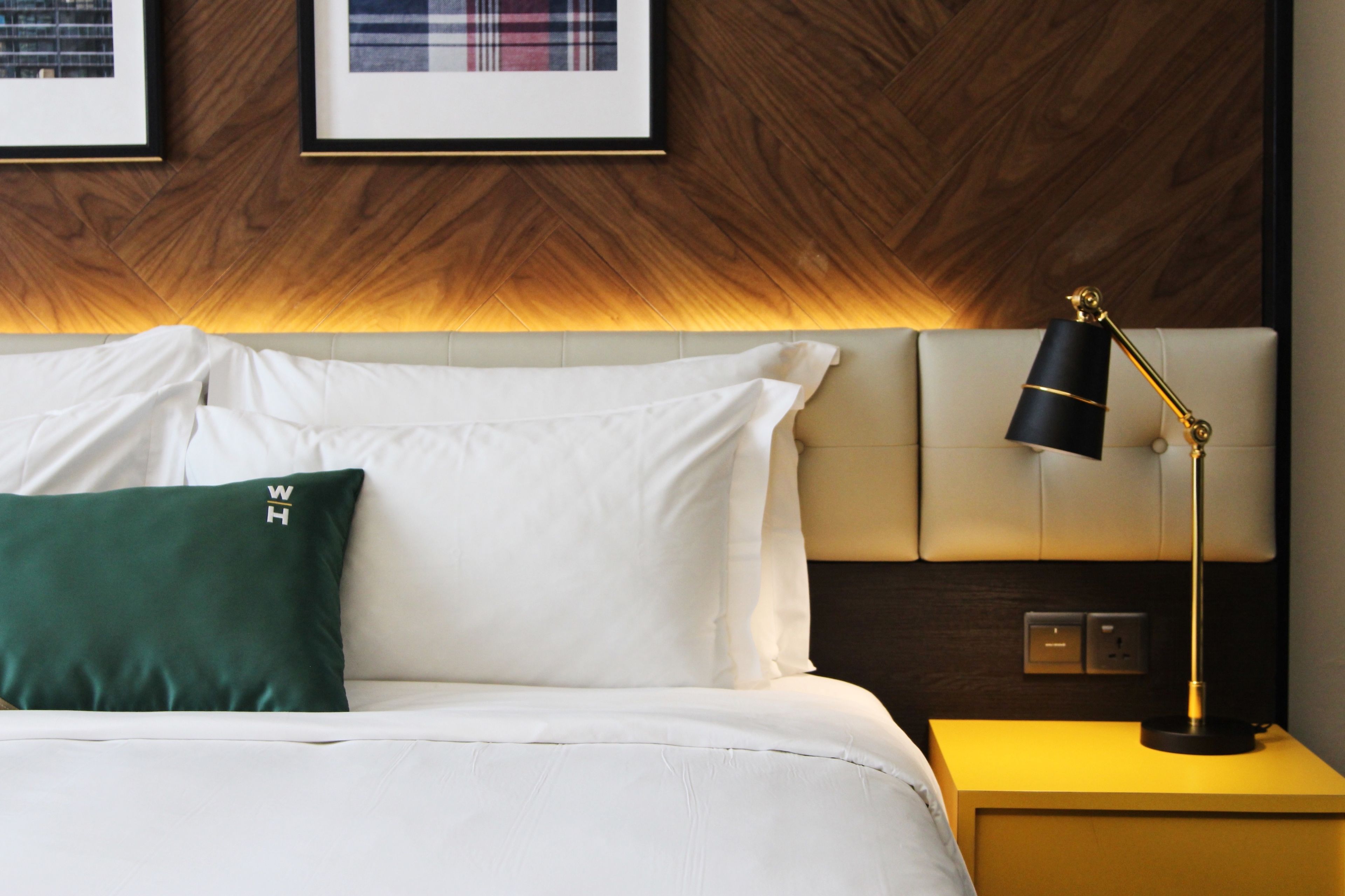 Una cama bien hecha con sábanas blancas y almohada verde, una lámpara de noche y cuadros en la pared.